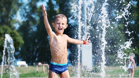 excited boy of seven years having fun between water jets, in fountain, run around, sprinkle, have fun, have fun, on a hot summer day. Summer in the city. Slow motion. High quality photoの写真素材