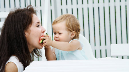 summer, in the garden, a funny one-year-old blonde girl treats her mother with watermelon, feeds her from her hands, the girl also eats watermelon. High quality photoの写真素材