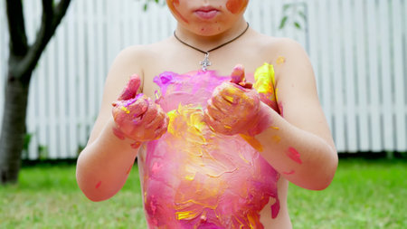 a little child, a four-year-old boy playing, painting with finger paints, decorating himself, in the garden, sitting on a blanket, on grass, lawn, in the summer. hes having fun. High quality photoの写真素材