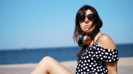 portrait, beautiful brunette, woman in sun glasses, and headphones, sitting on the beach, against...