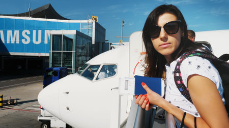 brunette woman in sun glasses, with backpack, holding Airline boarding pass tickets, and passport at airport, on background of the aircraft. summer. High quality photoの写真素材