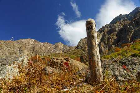 An old milepost stands guard on a dirt path cutting through a vivid autumn-colored meadow, with majestic cliffs rising under a peaceful sky, epitomizing the untouched beauty of highland natureの写真素材