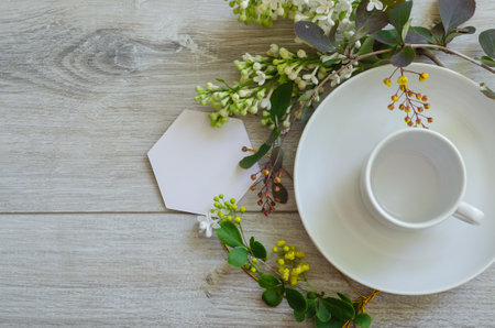 Flatlay with an empty cup on a saucer with mockup an empty white card framed by branches of green leaves, white lilac flowers and yellow small flowersの写真素材