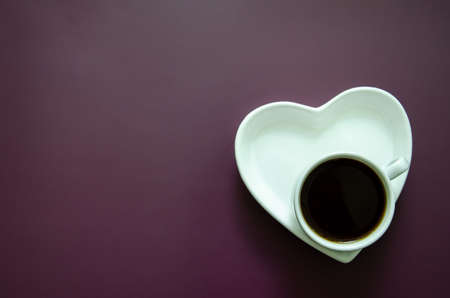 Flatlay A cup of coffee and a saucer in the shape of a heart in the lower corner, Flat lay white on a dark purple background, with large copyspaceの写真素材