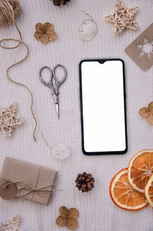 Wrapping gifts concept for holiday. Craft eco gift, dried orange slices, flowers, label, scissors, fir cones, rope on rose background. Smartphone top view mock up.の写真素材