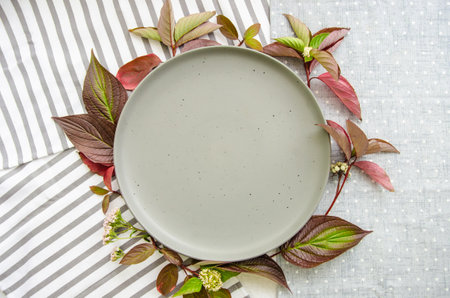 Ceramic plate with green and red plant leaves and wild flowers on cotton tablecloth and napkins. Organic food mock up. Template for recipes or food menuの写真素材