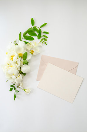Holiday greeting card mockup with envelope, blank card and white roses on white background, top view, flat lay. Blank wedding invitation card mockup and floral decorの写真素材