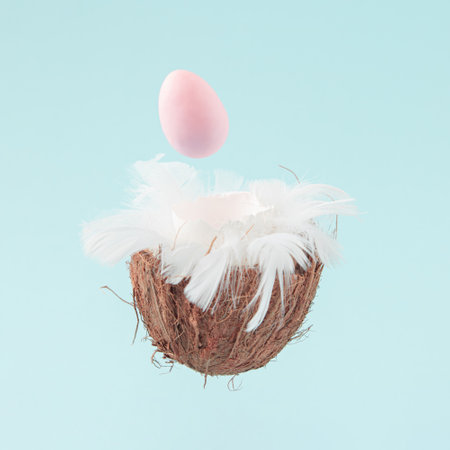 Easter pink egg with feathers in coconut shell shaped as a bird nest against pastel blue color. Creative minimal composition.の写真素材