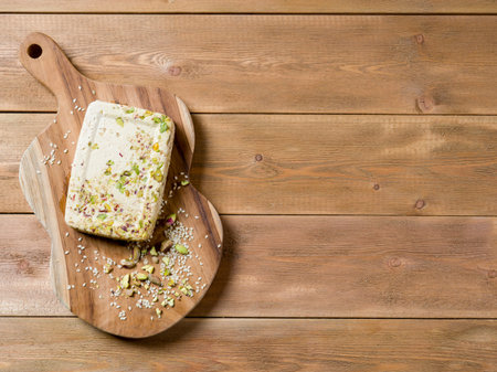 Sesame halva with pistachios on a teak chopping board on a brown wooden background. Top view. Copy...