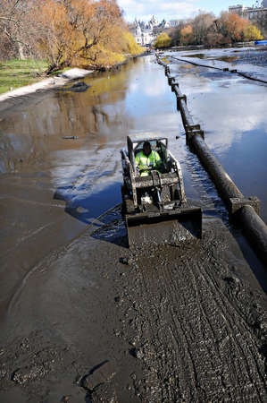 Worker in a small bulldozer excavating canal. Mud, pipeline, sky reflection in the waterの写真素材