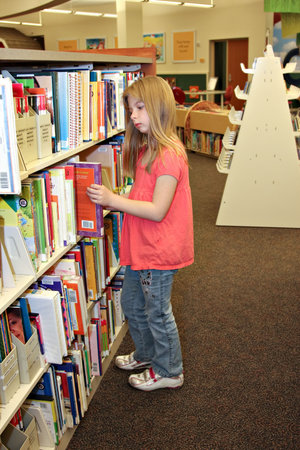 A school agied girl looks at books on bookshelf in library.