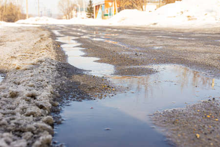 spring streams flow along the road covered with ice and heavy snow, the sky with clouds reflects in puddles, selective focusの写真素材