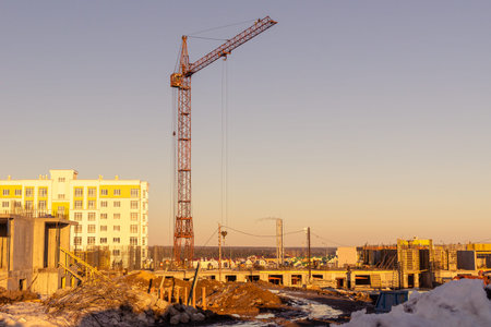 In the spring evening, a tower crane builds buildings on the outskirts of the city, lifting and...