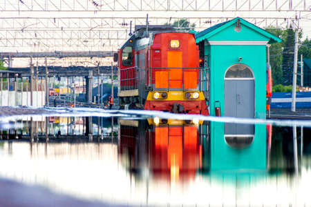Kemerovo, Russia, 23 June 2021 - red diesel locomotive at station is reflected in huge puddle on platform formed from mistakes made in designing the surface of the passenger platform, selective focusのeditorial素材
