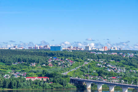 Kemerovo, Russia - June 24, 2021. Suburban village houses on shore and multi-storey residential buildings behind a forest on a hill, in foreground a road bridge across the Tom River, selective focusのeditorial素材