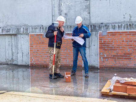 Kemerovo, Russia - 24 June 2021. Two engineers in hard hats are studying plan of structure while standing on wet concrete surface, in hands of one man is satellite geodetic instrument, selective focusのeditorial素材
