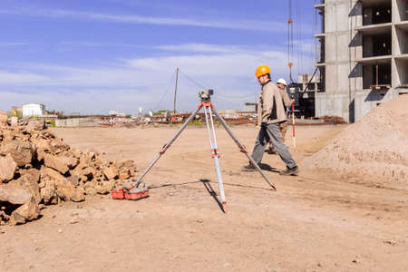 Kemerovo, Russia - Jule 02 2021. Optical level is installed on construction site near pile of large stone, worker in helmet passes by, surveyor works with satellite geodetic receiver, selective focusのeditorial素材