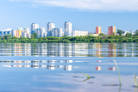 blue-white and red-yellow high-rise buildings behind coastal vegetation are reflected in the water of a lake or river, selective focusの写真素材