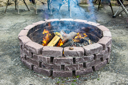A fire is burning in a round stone fire pit made of gray blocks, surrounded by grass and garden...