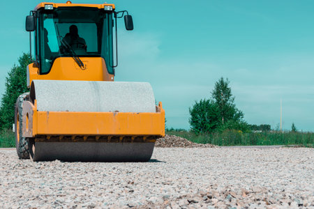 Machine with driver inside rolls on gravel making it firm and even while green plants and blue sky...