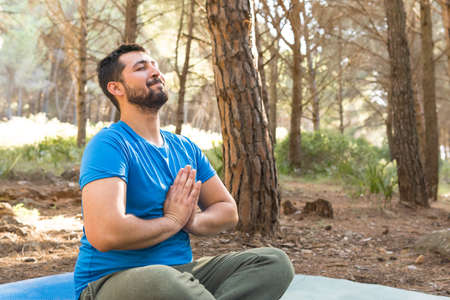 Handsome young adult relaxing practicing meditation sitting on his blue mat in nature at sunset surrounded by trees.の写真素材