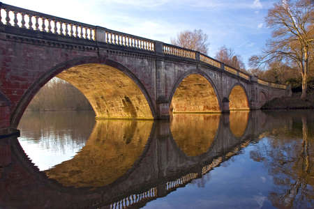 Sunlit arches of a stone & brick bridge, reflecting in a calm lake.の写真素材