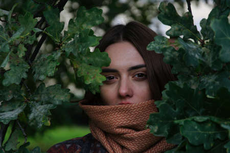 beautiful young woman with red hair looks out from behind the leaves of a tree in the park in autumnの写真素材
