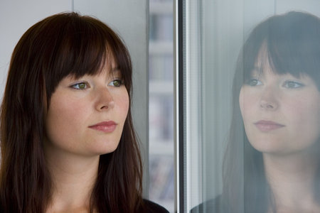 A beautiful young woman stares wistfully into a window where her reflection looks back at her.