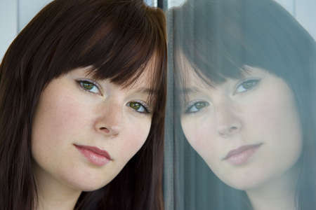 A beautiful young woman stares wistfully into a window where her reflection looks back at her.の写真素材