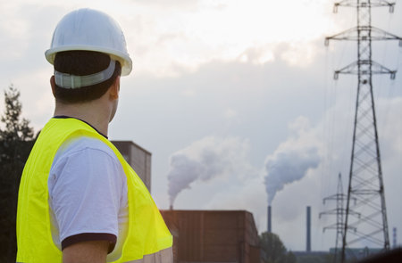 A man dressed in hardhat and safety hi-visibilty jacket surveys a factory with chimneys putting out...