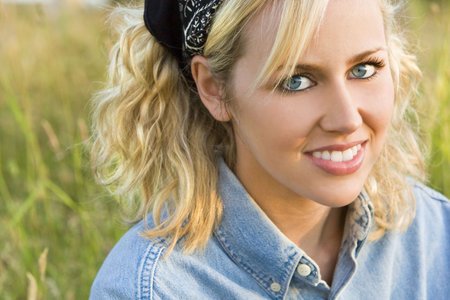 A beautiful blond haired blue eyed model wearing a black headscarf sits amid tall grass illuminated by golden evening sunlightの写真素材