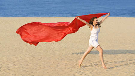 A sexy and beautiful young brunette woman running along a golden beach with a length of red material and a deep blue sea behind her.の写真素材