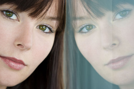 A beautiful young woman thoughtfully rests her head on a window where her reflection looks back.