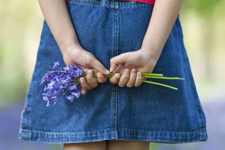 Shot of a little girl holding a bunch of blue bells behind her back, shot against an out of focus background of a bluebell wood.の写真素材