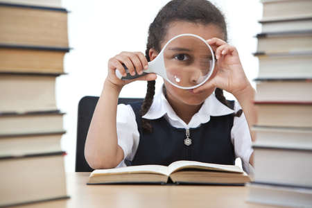 A beautiful young mixed race girl looking through a magnifying glass while reading in a school classroom with two piles of books in front of her.の写真素材