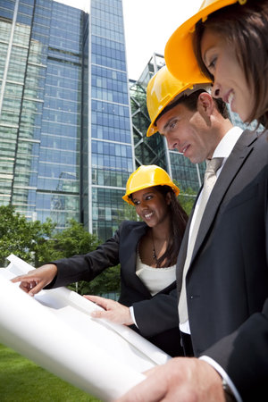A group of three executives, one man and two women, wearing hard hats review architectural plans in...