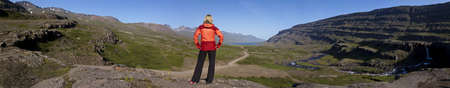 Panoramic shot of a woman hiker looking down the Berufjordur Valley in Iceland. The Berufjr<eth>ur fjord is visible at the end of the valley and a small waterfall can be seen on the right.の写真素材