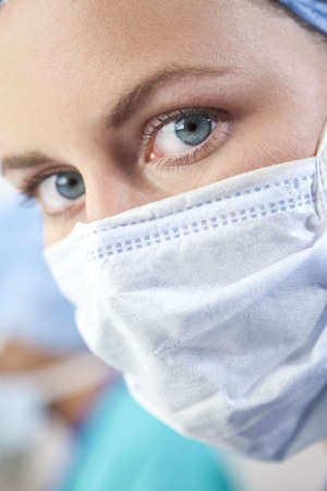 Close up portrait of a blue eyed female doctor wearing scrubs and a surgical mask, her colleague is out of focus in the distant background.の写真素材