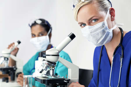 A blond female medical or scientific researcher or doctor using her microscope in a laboratory with her Asian colleague out of focus behind her.の写真素材