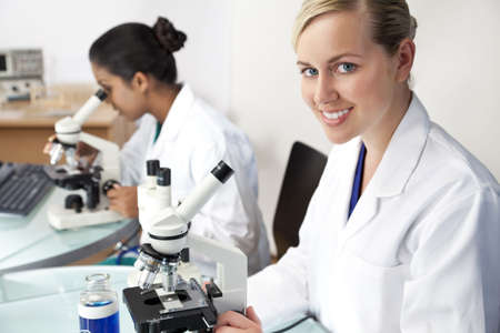 A blond female medical or scientific researcher or doctor using her microscope in a laboratory with her Asian colleague out of focus behind her.の写真素材