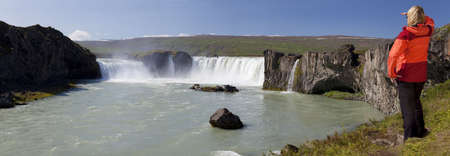 Panoramic shot of a young woman hiker looking at a waterfall. Shot on location at Godafoss waterfall in Iceland, the focus is on the waterfall.の写真素材