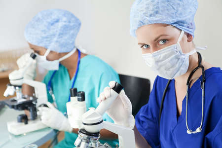 A blond female medical or scientific researcher or doctor using her microscope in a laboratory with her Asian colleague out of focus behind her.の写真素材