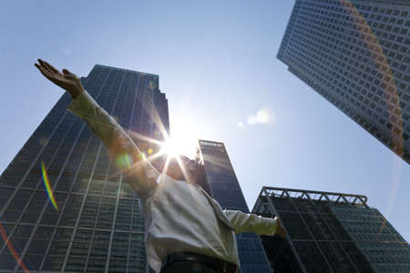 A businessman with his arms out stretched 'messiah like' in a modern city environment with the sun bursting over glass fronted office buldings behind himの写真素材