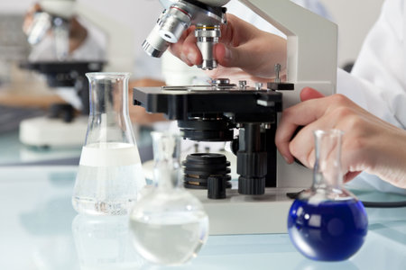 A medical or scientific researcher using a microscope in a laboratory environment with flasks and...
