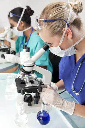 A blond female medical or scientific researcher or doctor using her microscope in a laboratory with her Asian colleague out of focus behind her.の写真素材