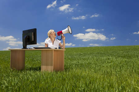 Business concept shot of a beautiful young woman sitting at a desk using a megaphone in a green field with a bright blue sky. Shot on location.の写真素材