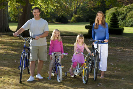 A young family with mother father and two blond daughters riding their bikes in a sun bathed green parkの写真素材