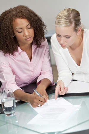 Two beautiful young businesswomen or lawyers, one African American one caucasian, discussing and signing a contract or legal documentの写真素材