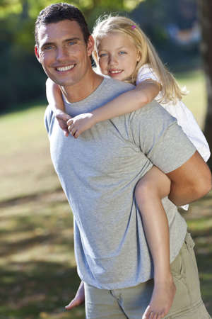 A young father with his blond daughter on his shoulders having fun in a sun bathed green parkの写真素材