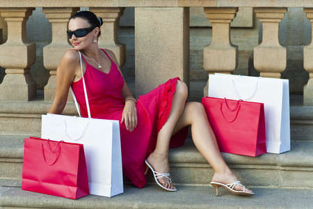 A classically beautiful latina woman in a red dress sitting with red and white shopping bags and looking happyの写真素材